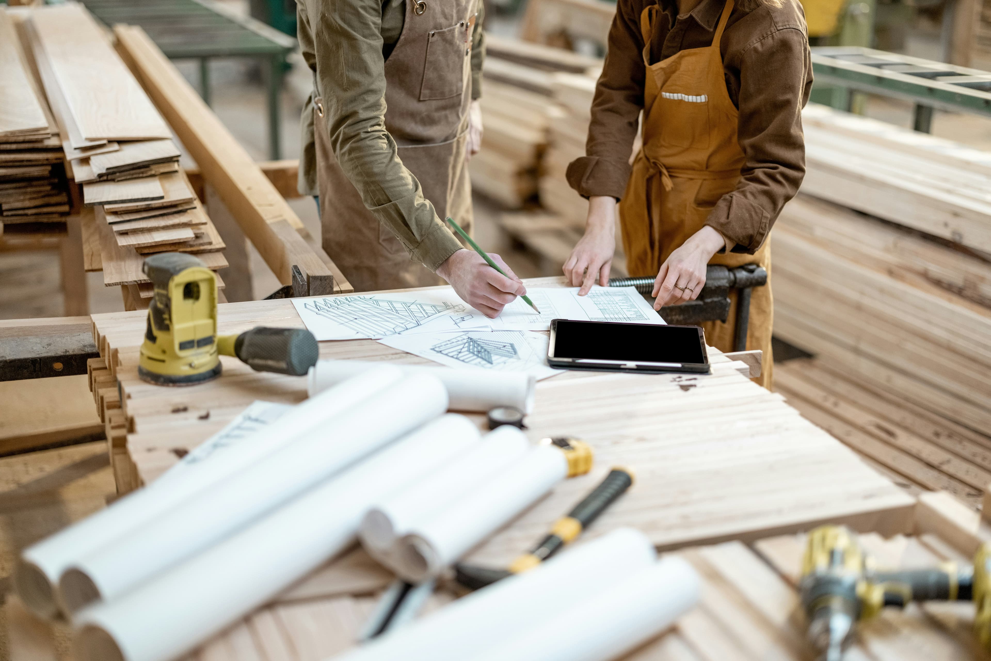 Two woodworkers in aprons review blueprints and a tablet on a workbench in a timber workshop.