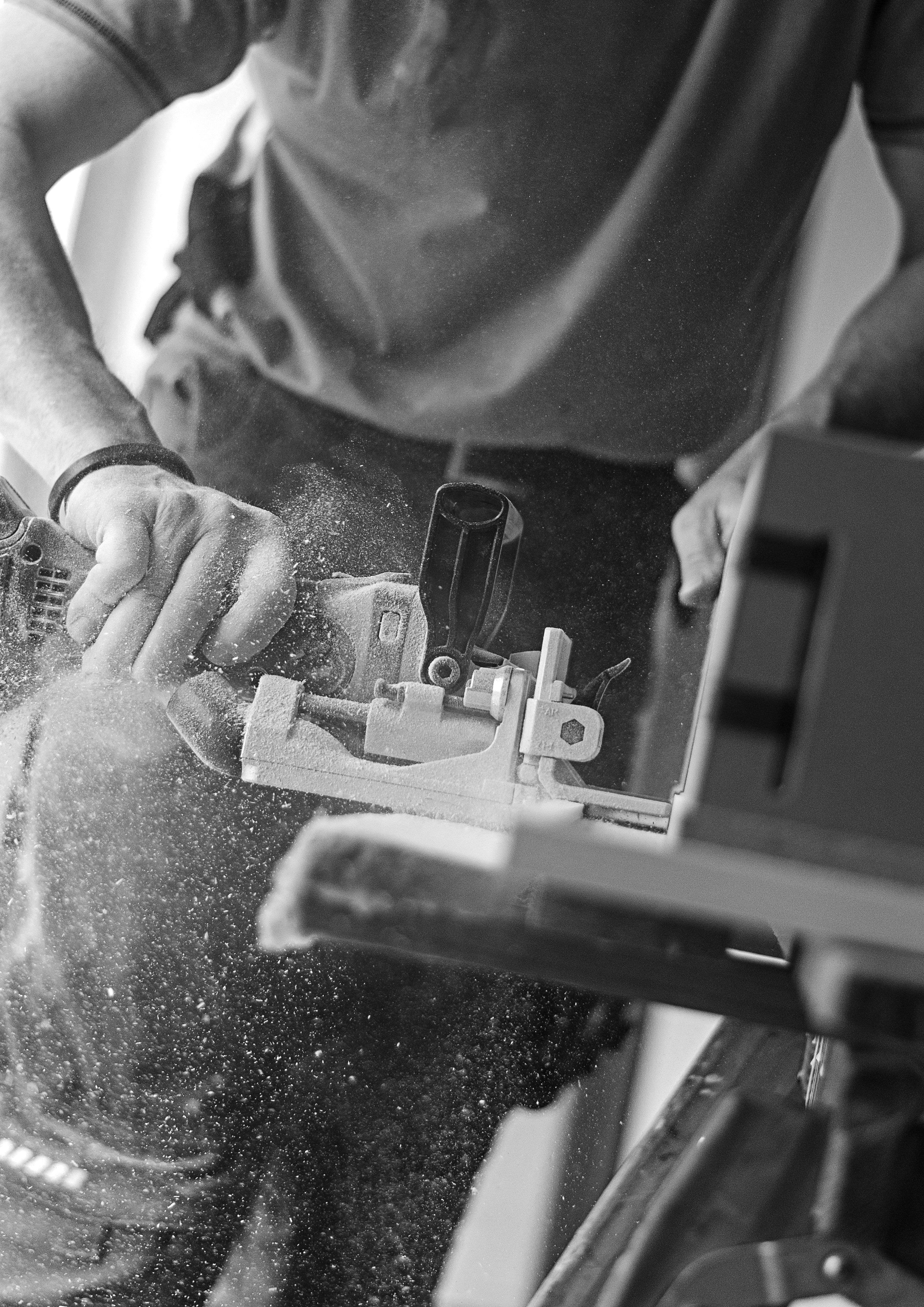A person uses a power tool to cut wood, sending sawdust flying, in black and white.